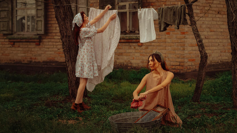 Two women performing a vintage-inspired outdoor laundry scene beside a brick building