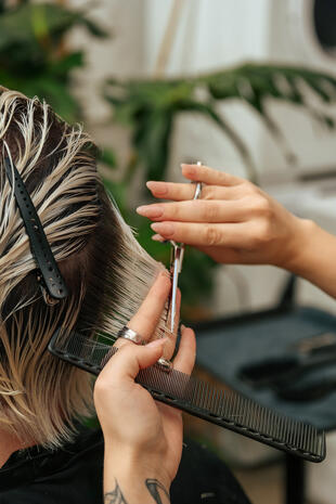 Close-up of a hairstylist’s hands cutting wet blonde hair with scissors and a black comb.