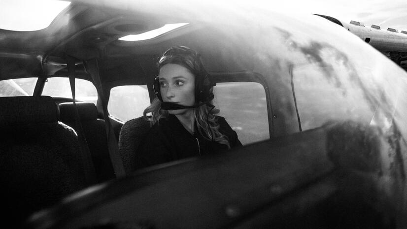 Woman wearing an aviation headset inside a small aircraft, photographed through the cockpit window