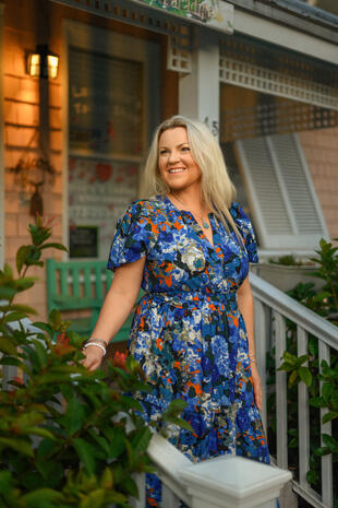 Smiling blonde woman in a blue floral dress standing outdoors beside a porch