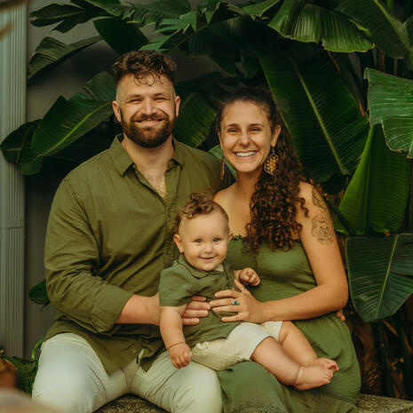 Two smiling parents hold their young child on their laps, wearing coordinated green outfits against large tropical leaves.