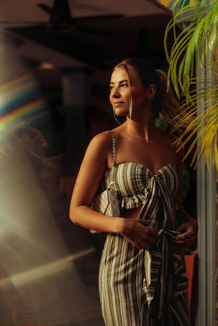 A woman in a striped outfit looks to the side beside palm leaves, with warm sunlight on her face and rainbow flare across the foreground.