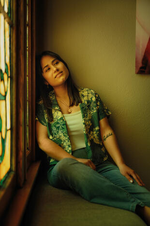 A woman in a green floral shirt and jeans sits beside a stained-glass window, resting her head against its wooden frame.