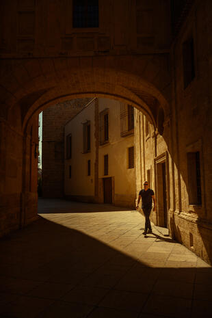 A man in dark clothing and sunglasses walks beneath a large stone arch, with warm sunlight casting long shadows across the pavement.