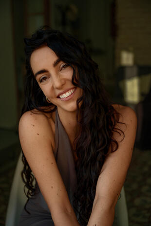 Close portrait of a smiling woman with long dark curls, leaning forward and looking at the camera against a muted green background.