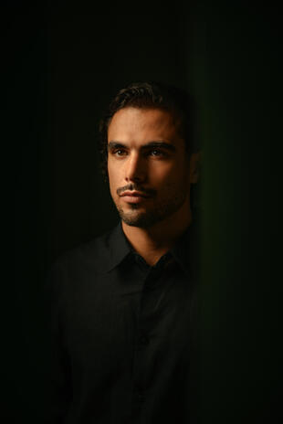 A man in a black shirt looks slightly away from the camera, his face lit from one side against a dark green background.