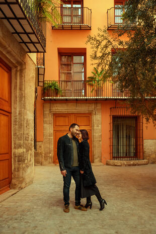 A couple leans close together in a courtyard framed by orange walls, wooden doors and wrought-iron balconies.