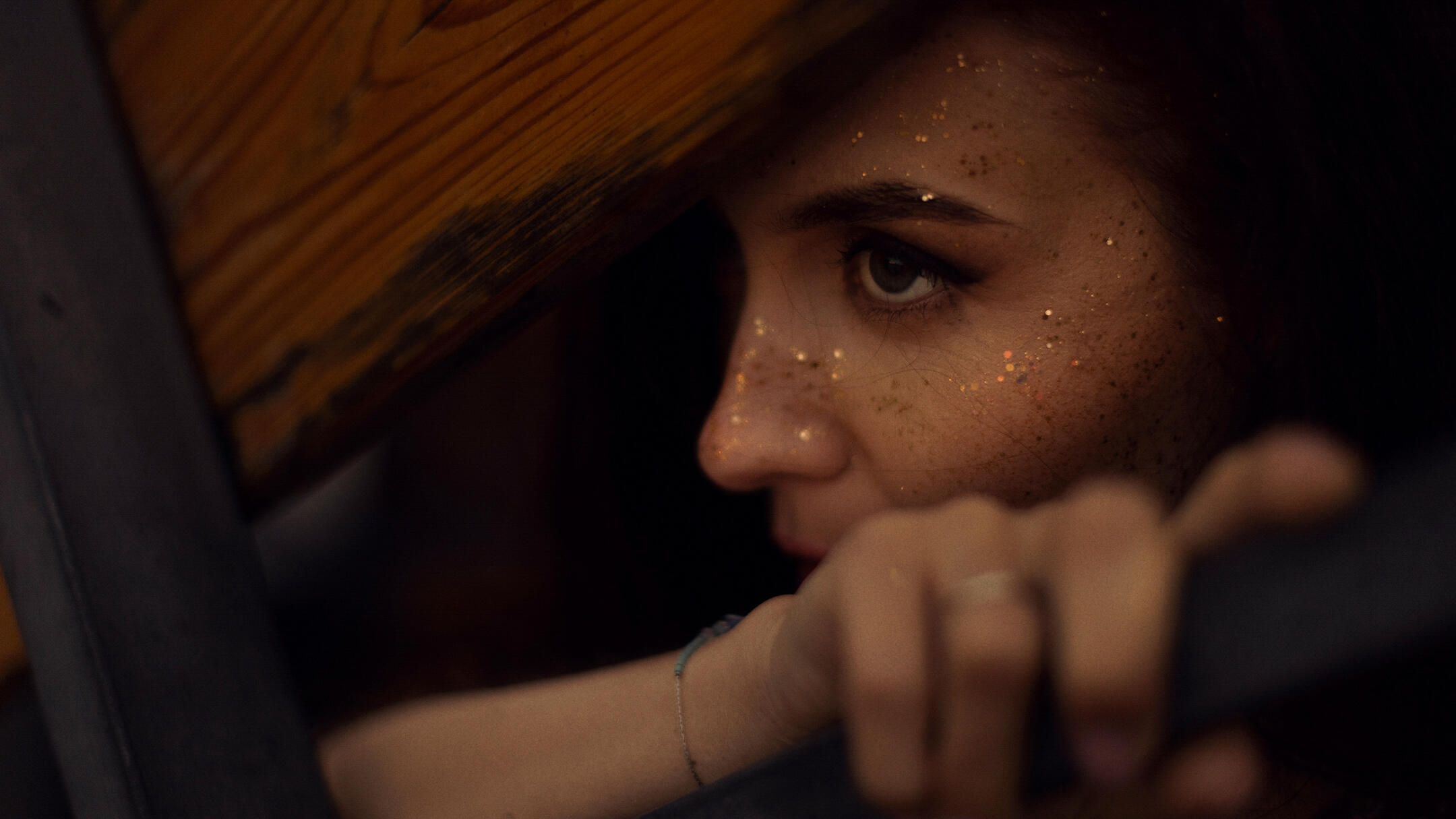 A portrait shaped by light and imagination Cinematic close portrait of a woman with gold-painted freckles looking through a dark wooden structure